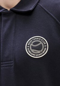 Μπλε ναυτικό πόλο με υφασμάτινη υφή, που διαθέτει στρογγυλό κεντημένο λογότυπο με τη φράση "BOSS TENNIS CLUB" και σχέδιο με μπάλα του τένις.