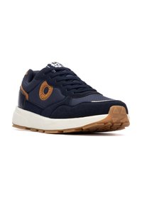 Zapatilla deportiva azul marino con materiales de ante y malla, con un detalle en cuero marrón, suela texturizada y media suela blanca. Detalle de logo circular.