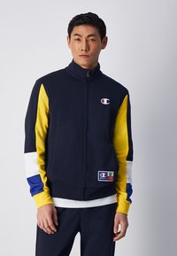 Giacca zip-up blu navy con pannelli gialli, bianchi e blu. Presenta un logo sul petto e un tessuto testurizzato con una tasca.