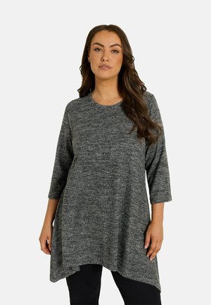 Vrouw met lang donker haar, die een losse grijze driekwart mouw top en zwarte broek draagt tegen een effen lichte achtergrond.