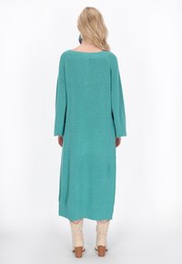 Abito in maglia turchese, di lunghezza media, con maniche ampie e scollatura a barca. Indossato con stivali di colore chiaro e tessuto strutturato visibile.
