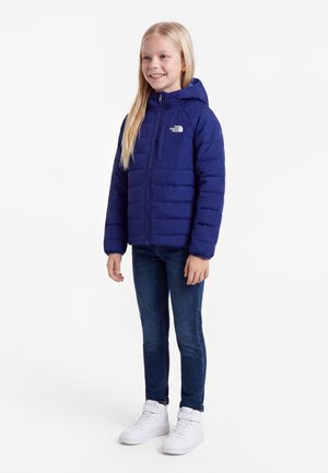 Smilende pige iført en blå hættejakke, mørke jeans og hvide sneakers, stående mod en enkel hvid baggrund.