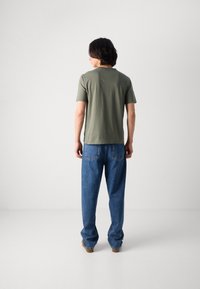 T-shirt en coton vert olive à manches courtes, associé à un jean en denim bleu clair avec des poches arrière, et des chaussures marron. Fond neutre.