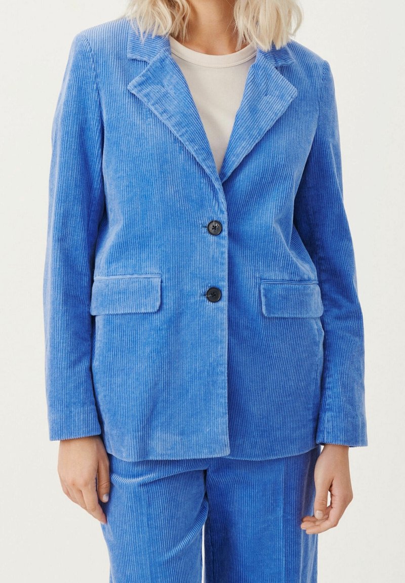 Femme portant un costume en velours côtelé bleu clair avec des boutons noirs, des poches à rabat et un sous-shirt blanc, montrée des épaules jusqu'à mi-cuisse.