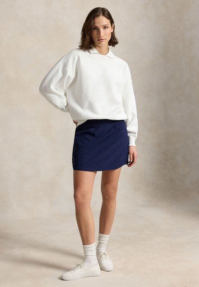 Femme aux cheveux courts châtains portant une chemise blanche à manches longues, une jupe bleu marine, des chaussettes blanches et des baskets blanches, debout devant un fond uni.