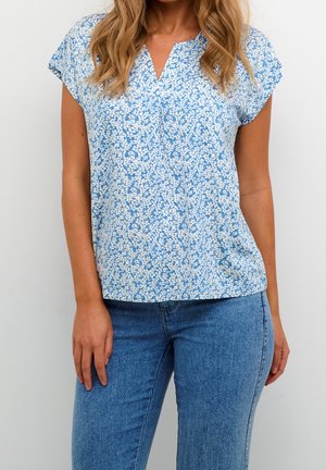 Kvinde iført kortærmet blå top med hvidt blomsterprint og blå jeans, stående mod en ensartet lys baggrund.