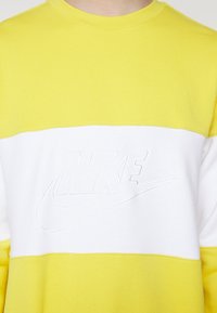 Žlutá a bílá color-block mikina s výšivkou bílého loga Nike na hrudi, zobrazená od krku po střed trupu.