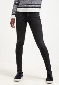 Jeans skinnies negros con una cintura de altura media, cinco bolsillos y una textura suave. Combinados con zapatos negros sin cordones y un suéter de patrón.