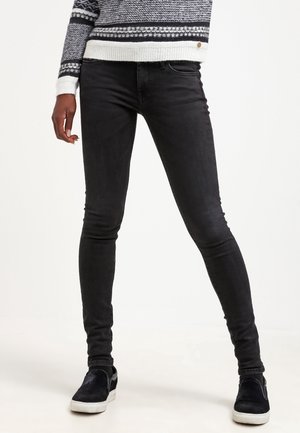 Jeans skinnies negros con una cintura de altura media, cinco bolsillos y una textura suave. Combinados con zapatos negros sin cordones y un suéter de patrón.