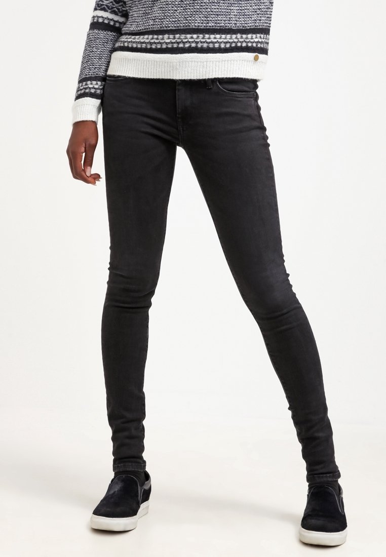 Jeans skinnies negros con una cintura de altura media, cinco bolsillos y una textura suave. Combinados con zapatos negros sin cordones y un suéter de patrón.