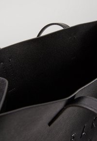 Czarna torba na zakupy z teksturowanym, gładkim wykończeniem. Posiada płaski top i wewnętrzne szwy, z delikatnie wytłoczonym logo z przodu.
