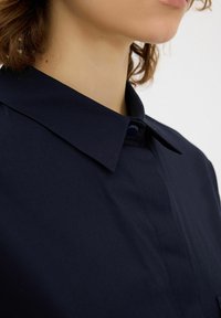 Námořnická modrá košile s límečkem má zapínání na knoflíky a hladkou texturu. Límeček je špičatý a látka se zdá být lehká a pevná.