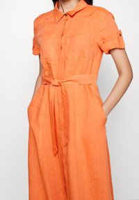 Oranžové linenové šaty košového střihu s krátkými rukávy, knoflíkovým límcem, dvěma náprsními kapsami a odpovídajícím páskem uvázaným v pase. Hladká textura.