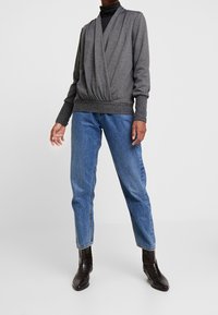 Szary sweter w stylu kopertowym z długimi rękawami, dekoltem w serek i prążkowanym brzegiem, zestawiony z niebieskimi, wysokim stanem jeansami oraz czarnymi botkami za kostkę.