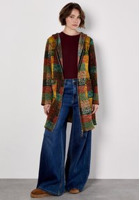 Vrouw in een multicolore patchwork capuchonjas, bordeaux overhemd, wijde blauwe jeans en bruine suède schoenen die tegen een witte muur staat.
