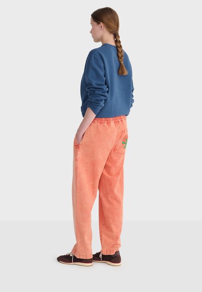 Femme aux cheveux tressés, vue de dos, portant un sweat-shirt bleu, un pantalon ample corail avec un motif de soleil vert sur la poche arrière, et des baskets foncées.
