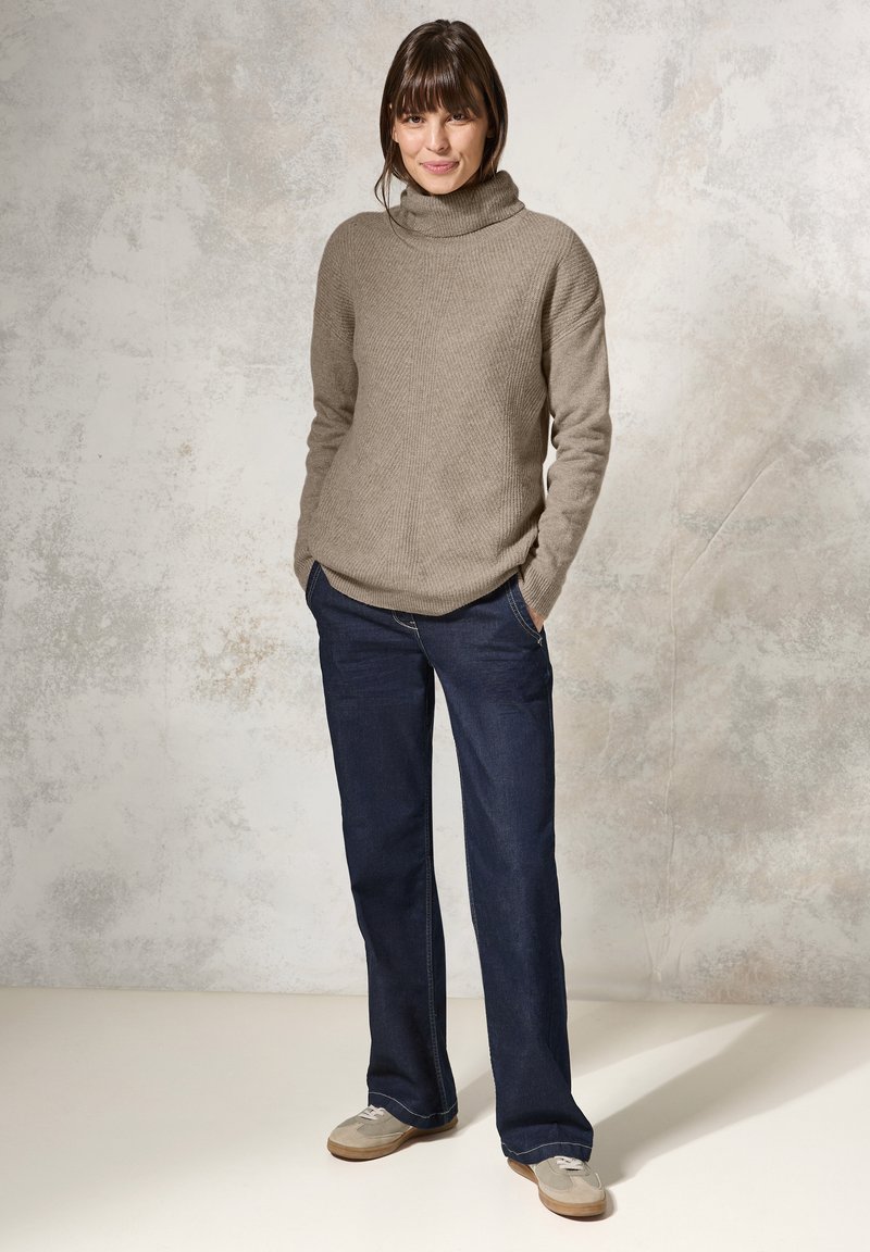 Beige geribbelde brei turtleneck trui gecombineerd met donkerblauwe wijde jeans en lichtgrijze suède schoenen. Gladde textuur en losse pasvorm.