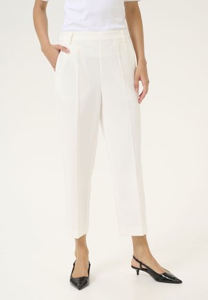 Femme portant un pantalon blanc sur mesure, longueur cheville, avec des plis sur le devant, associé à des escarpins noirs à bride arrière pointue.