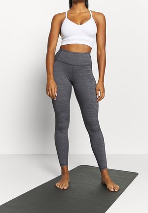 Leggings gris avec une finition texturée, assortis à un soutien-gorge de sport blanc arborant un logo sous les bras. Le mannequin est pieds nus sur un tapis de yoga.