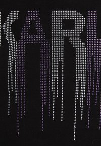 Svart tyg pryds med ordet "KARL" i stora rhinestones, med en gradienteffekt från silver till lila, vilket skapar ett droppande mönster.