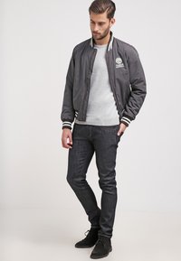 Grå bomberjacka med randiga ärmslut och krage, med en logotyp. Buren över en ljusgrå tröja, tillsammans med mörka jeans och stövlar.