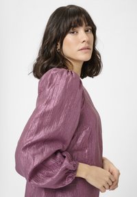 Frau mit schulterlangem, dunklem Haar, die eine glänzende, lila Bluse mit langen Puffärmeln trägt und vor einem schlichten weißen Hintergrund posiert.