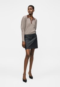 Γυναίκα ντυμένη με μπεζ ριγέ πόλο με μακρύ μανίκι, μαύρη μίνι φούστα και μαύρες μυτερές γόβες, στέκεται μπροστά σε λευκό απλό φόντο.