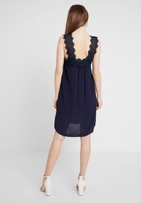 Kvinna i marinblå ärmlös klänning med spetsdetalj på ryggen och vita högklackade sandaler, stående med ryggen vänd mot en enfärgad bakgrund.