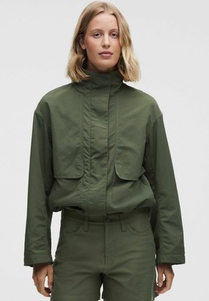 Frau mit blondem Haar, die eine olivgrüne Jacke mit hohem Kragen und passende Hosen trägt, steht vor einem schlichten hellen Hintergrund.