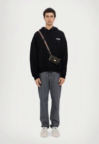 Sort hettegenser med frontlogo, kombinert med lyseblå jeans. En liten svart skulderveske med stripete stropp bæres. Hvite joggesko.