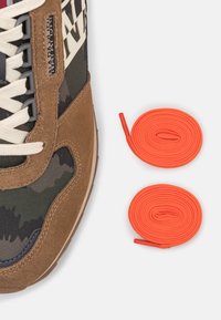 Sneaker in suede marrone con tessuto mimetico, lacci color crema e due matasse di lacci arancioni brillanti accanto. Texture liscia e forma solida.