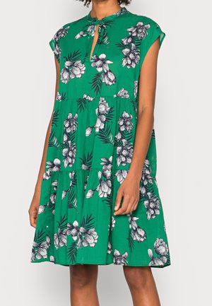 Zielona sukienka bez rękawów z czarno-białym kwiatowym wzorem, z falbanami na spódnicy i dekoltem z wycięciem, noszona przez osobę stojącą na białym tle.