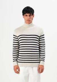 Uomo con un pullover a collo alto color crema con strisce orizzontali nere e pantaloni bianchi, in piedi davanti a uno sfondo bianco semplice.