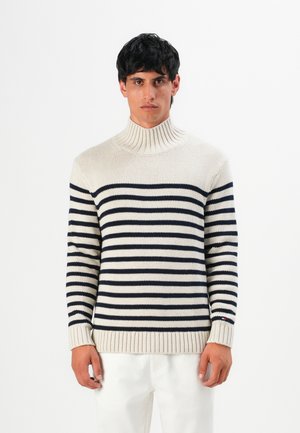 Mann, der einen cremefarbenen Rollkragenpullover mit schwarzen horizontalen Streifen und weißen Hosen trägt, vor einem schlichten weißen Hintergrund steht.