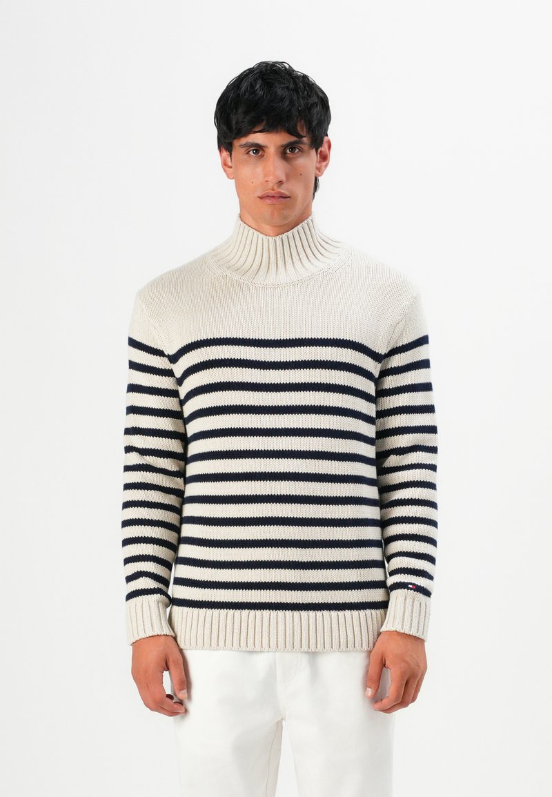 Uomo con un pullover a collo alto color crema con strisce orizzontali nere e pantaloni bianchi, in piedi davanti a uno sfondo bianco semplice.
