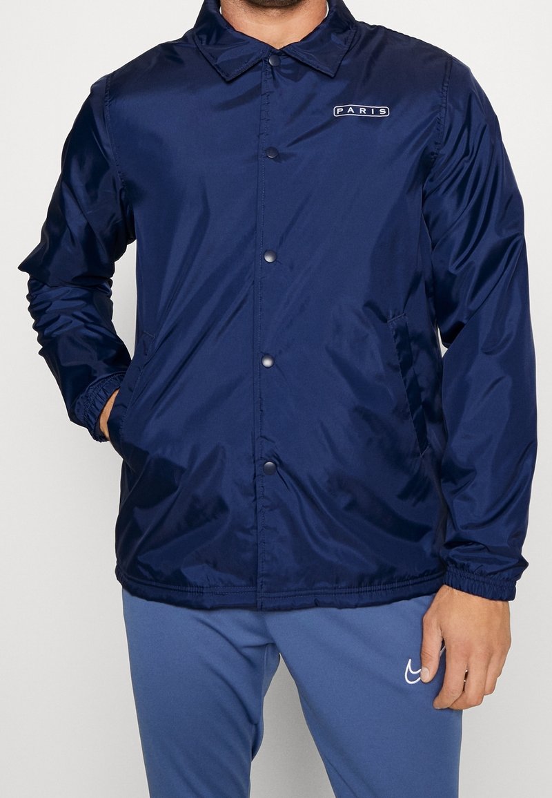 Giacca a vento blu navy in materiale lucido, con chiusura frontale a bottoni, polsini elasticizzati, tasche laterali e dettaglio logo "PARIS".