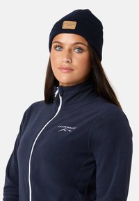 Tummansininen fleece-takki valkoisella vetoketjulla, jossa on logo. Yhdistetty tummansiniseen neulottuun pipoon, jossa on vaaleanruskea nahkapaikka. Sileä pinta.