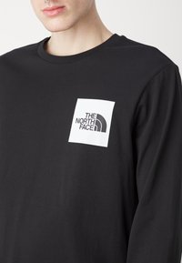 Schwarzes langärmliges T-Shirt mit einer weißen rechteckigen Logotasche, die den Text "THE NORTH FACE" und ein Bogen-Design in Schwarz zeigt. Glattes Baumwollgewebe.