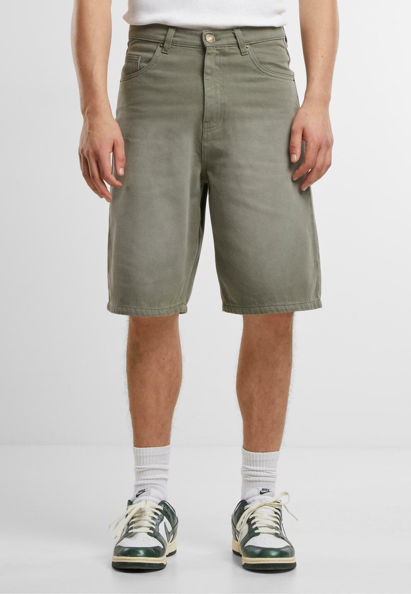 Shorts in denim verde oliva con taglio dritto, dotati di cinque tasche, passanti per la cintura e una texture liscia. Abbinati a sneaker verdi e bianche.