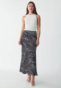 Schwarze Rock mit weißem Wellenmuster, gerippter weißer ärmelloser Oberteil und weiße Riemchen-High Heels, die eine fließende, moderne Silhouette schaffen.