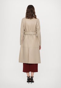 Bisas medvilninis ilgas paltas su diržu liemens srityje, struktūrine siluetu ir nugaros klostėmis, derinamas su raudonai rudos spalvos plačiomis kelnėmis ir juodais aukštakulniais sandalais.