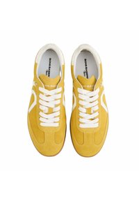 Des baskets en suède jaune présentent des lacets blancs et des accents latéraux, un bout arrondi et une semelle en caoutchouc beige. La marque est visible sur la semelle intérieure.
