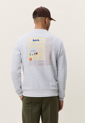 Homme portant un sweat-shirt gris clair avec un design coloré de devanture de café et un texte au dos, associé à un pantalon vert olive et une casquette marron.