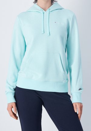Persoon die een lichtblauwe hoodie met voorzak en zwarte broek draagt, staand voor een effen achtergrond.
