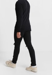 Chemise noire à manches longues associée à un jean noir déchiré et des baskets beige. La tenue présente une coupe ajustée avec une texture lisse.