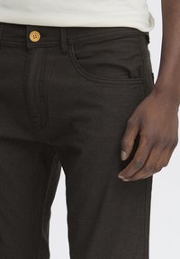 Črne jeans hlače slim fit kroja, s gumbom za zapiranje, sprednjimi žepe in diskretno teksturo. Roka počiva na tkanini.