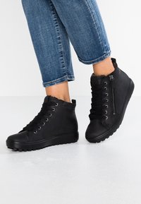 Sorte højskafter sneakers med snørebåndslukning og lynlås på siden, der har en glat overflade og gummisål. Bæres med opkrøllede denimjeans.