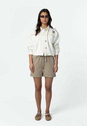 Kobieta w okularach przeciwsłonecznych, białej kurtce zapinanej na guziki z ozdobami, beżowych szortach i beżowych baletkach stojąca na białym tle.