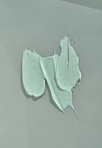 Texturerad mintgrön kräm med små bruna exfolierande partiklar, utbredd på en ljusgrå bakgrund, som visar en jämn och tjock konsistens.