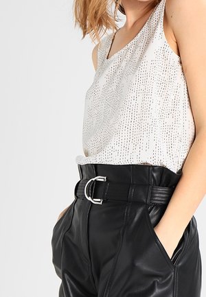 Kobieta w białym topie bez rękawów z wzorem, włożonym do wysokich czarnych skórzanych spodni z czarnym paskiem i srebrną klamrą.